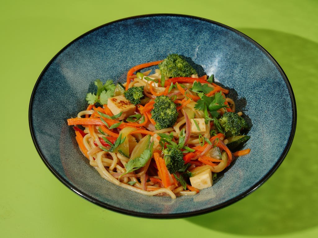 Vibrant noodle dish featuring tofu and fresh vegetables in a blue bowl against a green background.