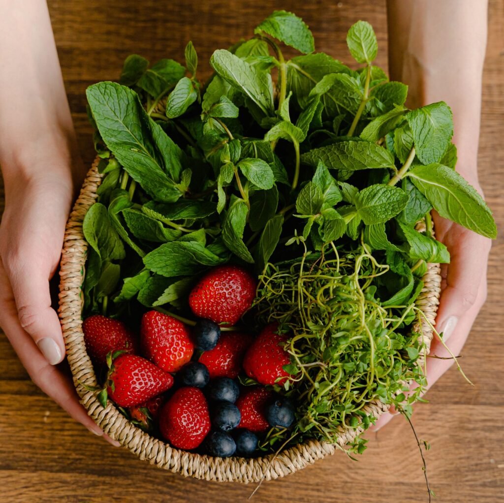 Top view of a basket filled with fresh strawberries, blueberries, and mint, held by hands on a wooden table.