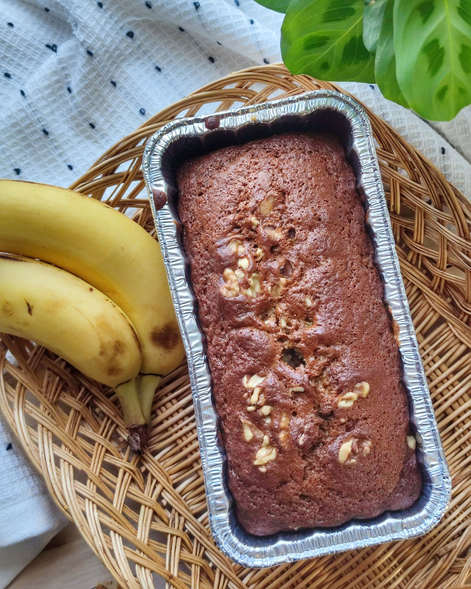 A freshly baked banana bread with walnuts, placed in a tin, paired with ripe bananas.