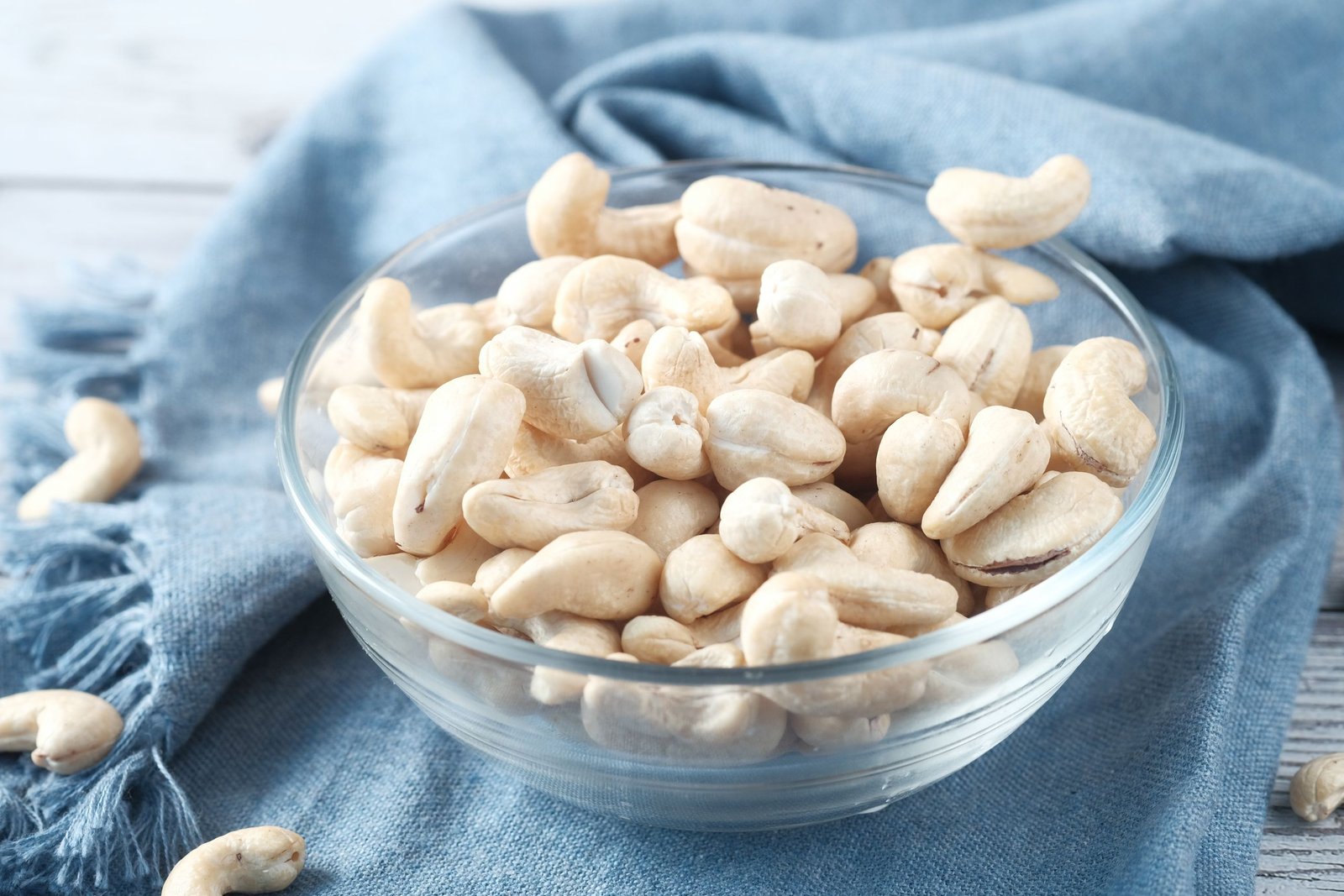 A glass bowl filled with raw cashews on a blue cloth, perfect for healthy snacking.