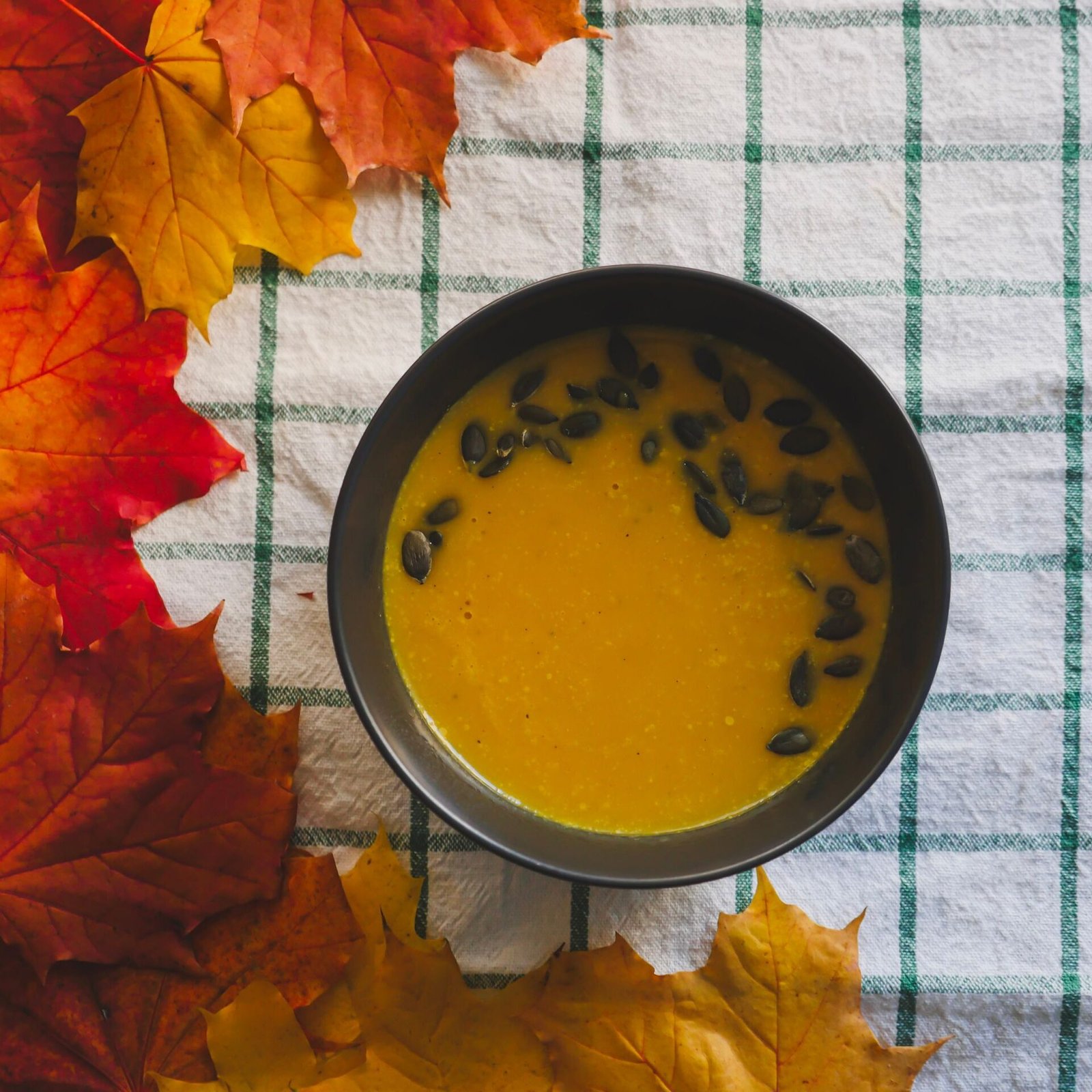Warm pumpkin soup in a bowl surrounded by vibrant autumn leaves on a checkered cloth.